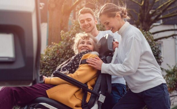 two helpers picking up disabled senior woman in wheelchair for transport