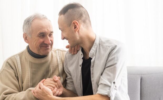 two generations male family old senior mature father and smiling young adult grown son enjoying talking chatting bonding relaxing having friendly positive conversation sit on sofa